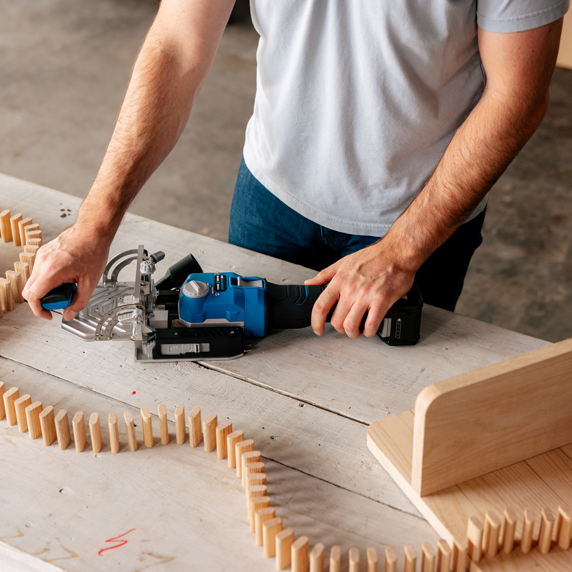 A man holds his Kreg Edge loose tenon joiner on a workbench, with several wooden tenons set up as dominoes curving around the power tool. 