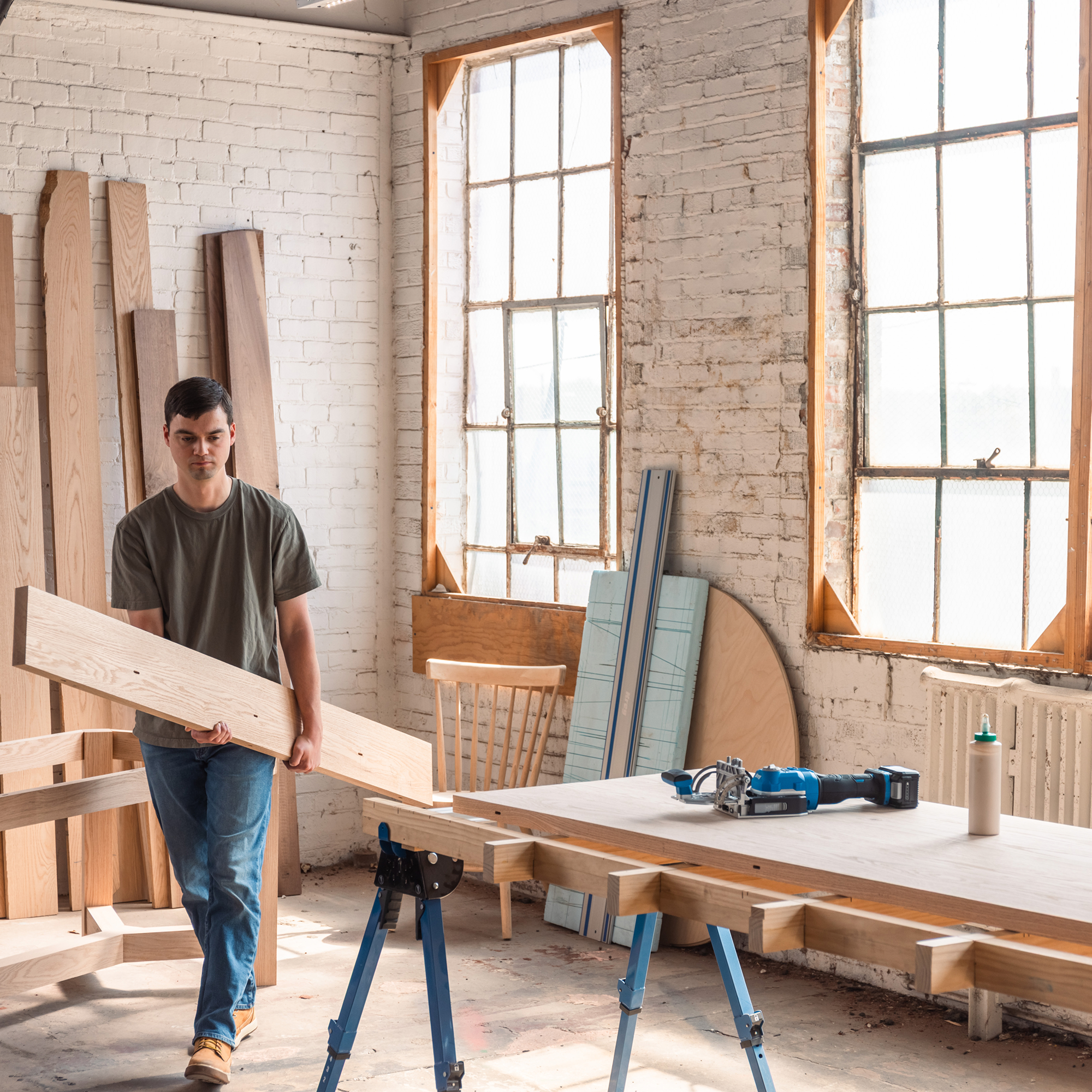 A man walks carrying a long wooden board. He carries it to his workspace in a warehouse where his Kreg Edge loose tenon joiner and a bottle of glue wait on the table.
