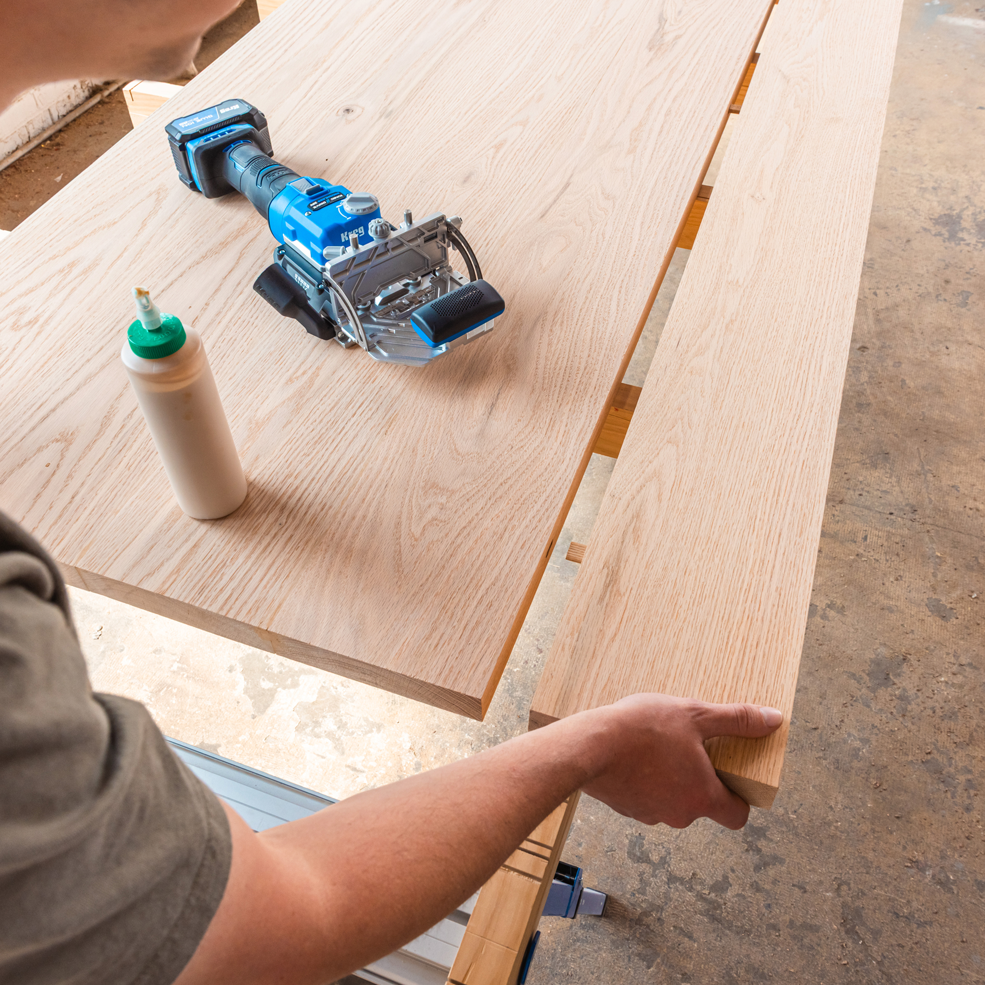 A hand is sliding a board closer to another board as tenons are connected with their mortises. The Kreg Edge loose tenon joiner and a glue bottle are on the table surface.