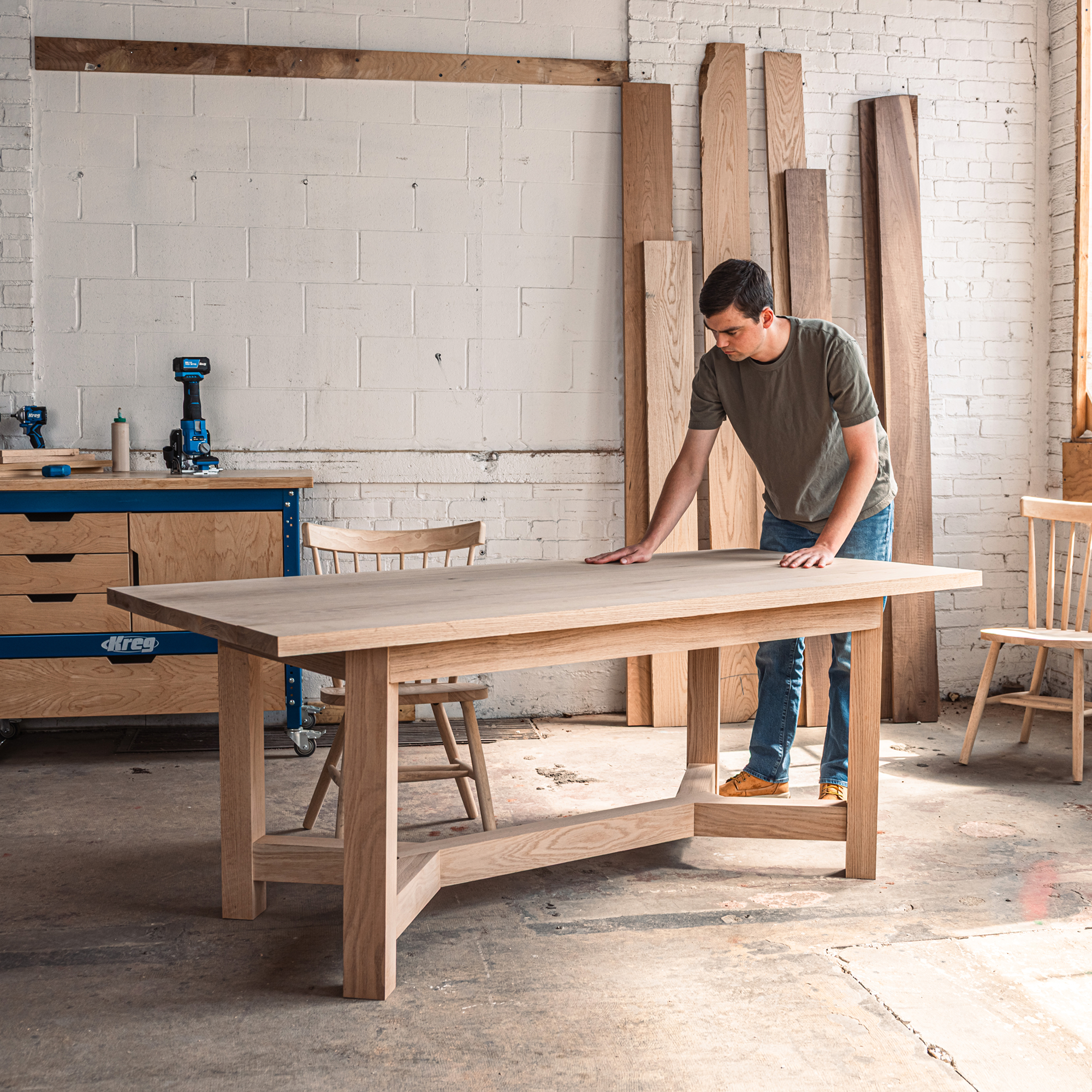 A man stands next to his newly built table in a workshop, inspecting his work.